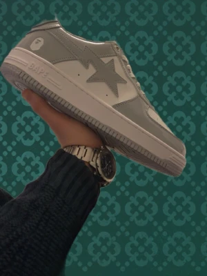 Gråvita BAPE STA sneakers - Snygga BAPE STA sneakers i grått och vitt med klassisk stjärndesign på sidan. Skorna har vita skosnören, perforerad tåbox och en robust vit sula. Tillverkade i blankt läder som ger en clean och modern vibe. Perfekta för dig som vill sticka ut med streetwear.