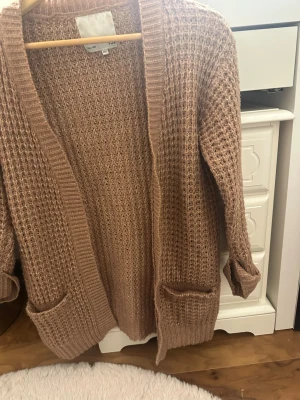 Beige stickad kofta med fickor - Mysig beige stickad kofta med grovstickat mönster och två stora fickor framtill. Koftan har lång ärm och öppen front utan knäppning, perfekt för lager-på-lager-stil. Ribbstickade kanter ger en snygg detalj.