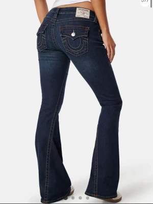 Mörkblå bootcut jeans True Religion - Säljer ett par mörkblå bootcut jeans från True Religion med kontrastsömmar och ikoniska fickdetaljer bak. Jeansen har låg midja och klassisk femficksdesign. Perfekta för dig som gillar en tidlös och snygg look. säljer då jag verkligen behöver pengarna