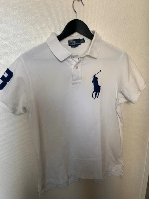 Vit pikétröja från Polo Ralph Lauren - Vit pikétröja från Polo Ralph Lauren med klassisk krage, knappar framtill och stor marinblå logga på bröstet. Tröjan har korta ärmar och en blå siffra på ena ärmen. Perfekt för en sportig och stilren look. Passar även S jätte bra. 