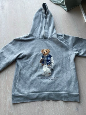 Grå hoodie Polo Bear Ralph Lauren - Grå hoodie från Ralph Lauren med ikoniska Polo Bear-trycket på bröstet. Björnen har på sig en blå och vit collegejacka och håller en amerikansk fotboll. Hoodien har huva och ribbade muddar. Perfekt för en avslappnad och cool stil. Tröjan passar allt från 155-163 cm och är xs