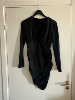 Svart omlottklänning med drapering - Snygg svart kort omlottklänning med lång ärm och draperad detalj på sidan. Djup v-ringning framtill och figurnära passform. Perfekt för dig som gillar stilrena och trendiga plagg.