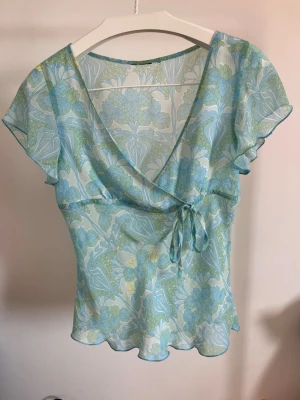 Blommig omlottblus i pastellfärger - Söt omlottblus med blommigt mönster i ljusblått, grönt och vitt. Blusen har korta, fladdriga ärmar och knytdetalj i sidan. Tunt, transparent material med vågig nederkant och djup v-ringning. Perfekt för en somrig look. 