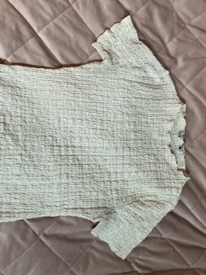 Vit ribbad topp med volangkant - Säljer en söt vit ribbad topp med korta ärmar och volangkant vid halsringning och ärmslut. Toppen har en stretchig, smockad struktur som ger en tight passform och en trendig look. Perfekt att styla med jeans eller kjol.