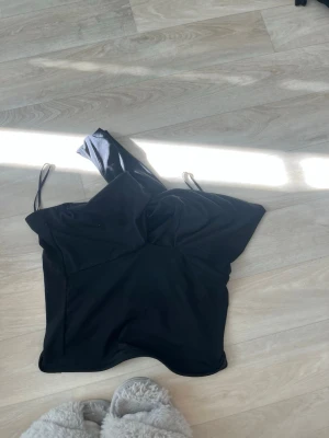 Svart axelbandslös topp - Snygg svart topp med tunna axelband och en axelbandslös design på ena sidan. Toppen har en croppad passform och är perfekt för att skapa en trendig look. Materialet känns mjukt och följsamt.