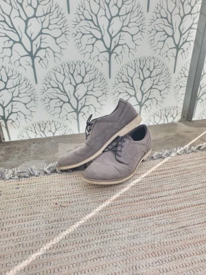 Gråa mockaloafers med snörning - Säljer ett par stilrena gråa loafers i mocka med snörning och ljus sula. Skorna har en klassisk, rundad tå och diskreta sömmar som ger en clean look. Perfekta för dig som gillar enkel men snygg stil.