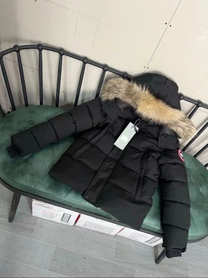 Svart dunjacka från Canada Goose - Svart dunjacka från Canada Goose med puffad form och stor huva med beige/brun pälskant. Jackan har logotyp på ärmen, dragkedja och knappar framtill. Materialet är polyester och dun, perfekt för vinter och riktigt kallt väder.