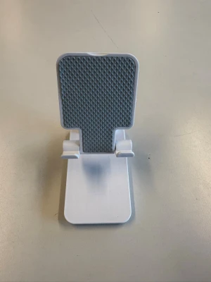 Vikbart mobilställ i vit plast - Snyggt och smidigt vikbart mobilställ i vit plast med grått mönstrat stöd. Perfekt för att hålla mobilen upprätt på skrivbordet när du kollar videos eller pluggar. Passar de flesta smartphones och är lätt att ta med tack vare den kompakta designen.
