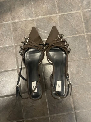 Bruna spetsiga pumps med remmar - Snygga bruna pumps med spetsig tå och flera tunna remmar med silverfärgade spännen. Skorna har en smal klack och öppen häl med justerbar ankelrem. Materialet ser ut att vara mockaimitation. Perfekta för att lyfta din outfit.