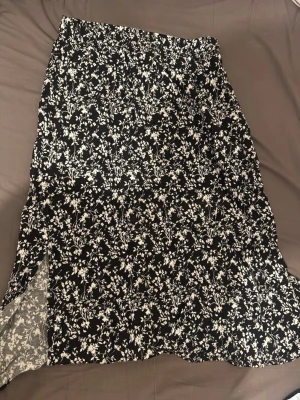 Svart långkjol med blommigt mönster - Svart långkjol med vitt blommigt mönster. Kjolen har en lätt utsvängd form och är perfekt för dig som gillar en flowy och bekväm stil. Passar snyggt till både sneakers och boots. Har även fickor och i väldigt bra skick.