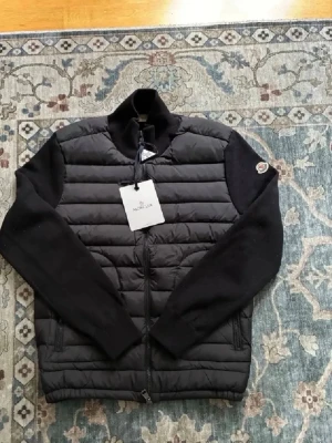 Svart cardiganjacka från Moncler - Snygg svart cardiganjacka från Moncler med quiltad front och stickade ärmar. Jackan har dragkedja framtill, ståkrage och två fickor med dragkedja. Moncler-logga på ärmen ger en exklusiv touch. Perfekt för lager-på-lager-stil.