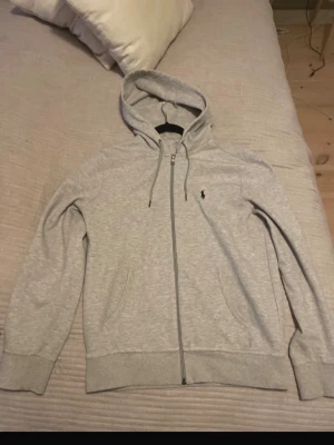 Grå hoodie från Polo Ralph Lauren - Snygg ljusgrå hoodie med dragkedja från Polo Ralph Lauren. Klassisk design med huva och dragsko, två fickor framtill och den ikoniska broderade loggan på bröstet. Perfekt för en avslappnad och stilren look.
