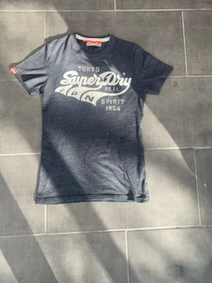 Mörkgrå Superdry t-shirt med tryck - Snygg mörkgrå t-shirt från Superdry med stort vitt tryck på bröstet. Klassisk rund hals och korta ärmar. Perfekt för en avslappnad och trendig stil. Materialet känns mjukt och skönt mot huden.