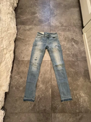 Ljusblå slitna skinny jeans - Säljer ett par ljusblå skinny jeans med slitningar och råa benslut. Jeansen har klassisk femficksdesign och dragkedjegylf. Perfekta för en avslappnad och trendig look.