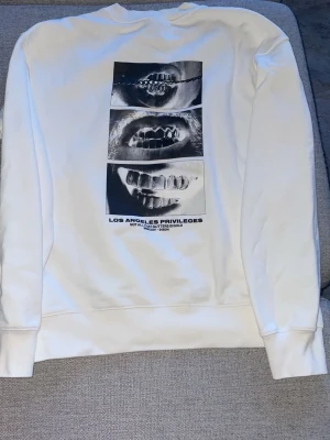 Vit sweatshirt Los Angeles Privileges - Vit sweatshirt med tryck av tre svartvita bilder på tänder på ryggen och liten text på bröstet. Tröjan har rund hals, långa ärmar och ribbade muddar. Materialet ser ut att vara mjukt bomullstyg. Perfekt för dig som gillar streetwear och grafiska prints.