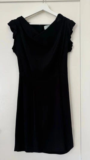 Snygg figursydd klänning med draperad framsida! - En supersnygg klänning för många tillfällen. Lätt drapering fram vilket gör den lite festligare. Dragkedja bak. Korta ärmar med volanger. Inga skavanker! Material är jag osäker på men gissar polyester och viskos. Axelmått 36 cm. Längd 86 cm.