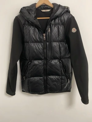 Moncler cardigan   - Snygg svart pufferjacka från Moncler med quiltad front och huva. Jackan har släta svarta ärmar, två dragkedjefickor fram och Moncler-logga på ena ärmen. Perfekt för kalla dagar och ger en clean, modern look. Kom privat för minsta fråga!