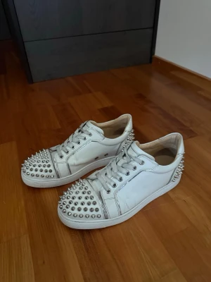 Vita sneakers med nitar från Christian Louboutin - Snygga vita sneakers i läder från Christian Louboutin med silverfärgade nitar på tån och hälen. Klassisk låg modell med vita snören och coola detaljer som sticker ut. Perfekta för dig som vill ha något unikt och edgy i garderoben. Låda finns, och kvitto! (Dom är äkta)