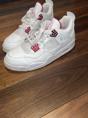 Vita Air Jordan 4 Retro sneakers - Snygga vita Air Jordan 4 Retro sneakers med röda detaljer och meshpaneler. Skorna har en klassisk siluett med högt skaft, vita skosnören och synlig Air-enhet i sulan. Tillverkade i läder och textil för en clean och sportig look.