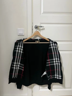 Burberry kofta - Snygg svart hoodie från Burberry med klassiskt rutigt foder i svart, vitt och rött. Dragkedja framtill och broderad Burberry-häst på bröstet. Perfekt för dig som vill ha en hoodie med exklusiv känsla och stilrena detaljer.