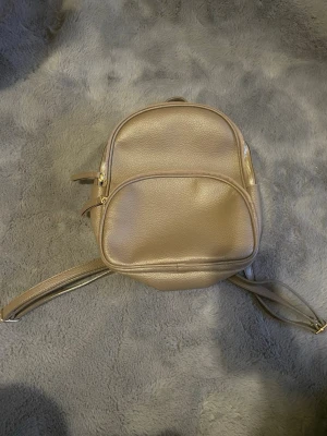 Beige miniryggsäck i skinnimitation - Snygg beige miniryggsäck i skinnimitation med justerbara axelremmar och guldfärgade dragkedjor. Väskan har ett stort huvudfack och en mindre ficka framtill, perfekt för att bära med sig det viktigaste. Enkel och stilren design som passar till många outfits.