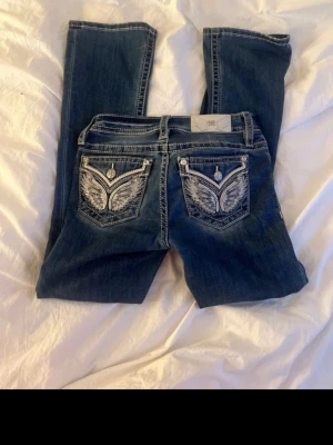 Blå jeans med broderade bakfickor - Snygga blå jeans från Miss Me med unika broderade detaljer och knappar på bakfickorna. Jeansen har klassisk femficksdesign och kontrastsömmar. Perfekta för dig som vill sticka ut med coola detaljer och en trendig look.