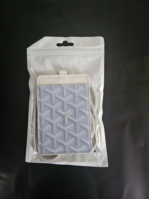 Ljusblå korthållare med snodd - Snygg ljusblå korthållare med geometriskt mönster och vita detaljer. Kommer med en matchande vit snodd med metallspänne, perfekt att hänga runt halsen. Praktiskt fönster på baksidan för ID eller busskort. Materialet är konstläder och textil.