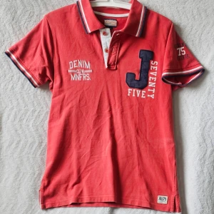 Röd pikétröja från Jack & Jones - Snygg röd pikétröja från Jack & Jones med vita och marinblå detaljer. Tröjan har korta ärmar, krage med knappar och broderade tryck på bröstet och ärmen. Perfekt till en avslappnad stil och passar till jeans eller shorts.