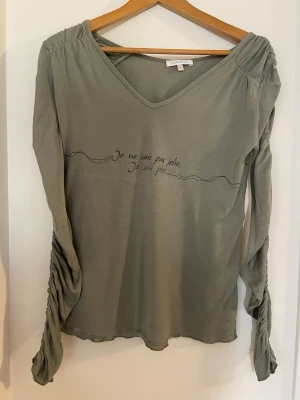 Grågrön långärmad topp från Kookai - Stilren grå långärmad topp från Kookai med v-ringning och rynkade detaljer längs ärmarna. Tryck på bröstet med fransk text och dekorativ linje både fram och bak. Tunn och mjuk kvalitet. Det finns ett pyttelitet hål i ärmen som jag inte märkt själv när jag köpte den som säkert kan fixa enkelt.