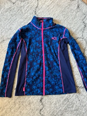 Blå mönstrad fleecejacka Kari Traa - Snygg blå fleecejacka från Kari Traa med blommigt mönster i olika blå nyanser och rosa detaljer. Jackan har hel dragkedja, hög krage och långa ärmar med rosa sömmar. Perfekt för lager-på-lager och chill dagar.