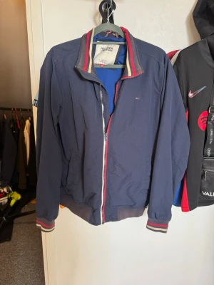 Blå vindjacka från Tommy Hilfiger - Snygg marinblå vindjacka från Tommy Hilfiger med dragkedja och ribbade muddar. Jackan har Hilfigers klassiska ränder på krage och ärmslut samt diskret logga på bröstet. Perfekt för dig som gillar stilrent
