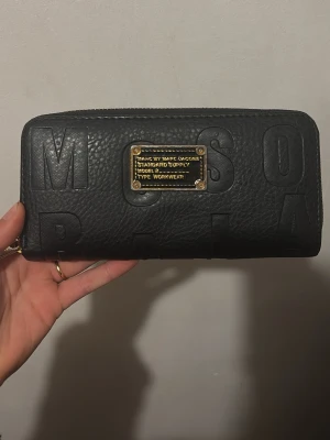 Svart plånbok från Marc Jacobs - Cool svart plånbok från Marc Jacobs med präglad logotyp och guldfärgad metallplatta på framsidan. Plånboken är tillverkad i läder och har flera fack för kort, sedlar och mynt samt ett snyggt mönstrat innerfoder. Dragkedja runt om för extra säkerhet. Otroligt praktisk och bärbar. 