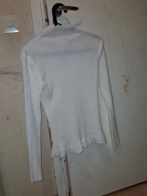 Vit ribbad polotröja (För kvinnor  - Säljer en vit ribbad polotröja med långa ärmar och figurnära passform. Tröjan har en hög hals och vågig kant längst ner, vilket ger en snygg detalj. Perfekt att styla med jeans eller kjol för en clean look.