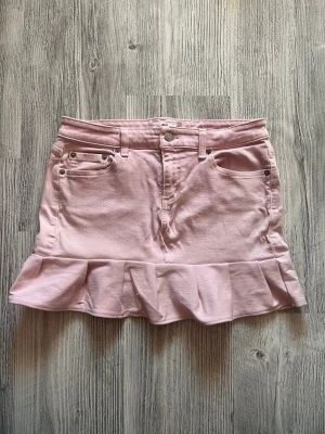 Ljusrosa volangkjol i denim - Supersöt ljusrosa kortkjol i denim med volangkant nedtill och klassiska jeansfickor både fram och bak. Kjolen har bälteshällor och stängs med knapp och dragkedja. Perfekt för dig som vill ha en feminin och trendig look.