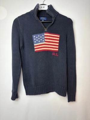 Mörkblå stickad tröja Polo Ralph Lauren - Mörkblå stickad tröja med halv dragkedja från Polo Ralph Lauren. På bröstet finns en stor amerikansk flagga i rött, vitt och blått samt RL-broderi. Tröjan har ribbstickade muddar och hög krage. Perfekt för dig som gillar klassisk college-stil. Storlek XL i barn passar XS