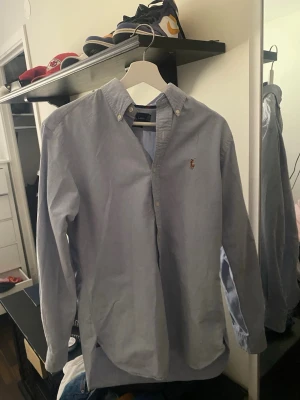 Ljusblå skjorta från Ralph Lauren - Klassisk ljusblå skjorta från Ralph Lauren med button-down krage och broderad logga på bröstet. Skjortan har långa ärmar och knäppning framtill. Perfekt för en stilren och avslappnad look.