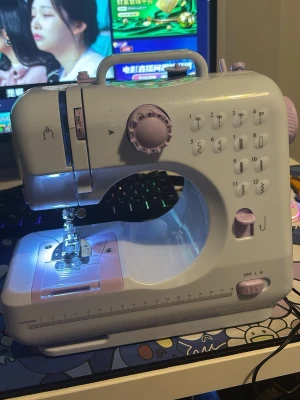 Ljuslila symaskin med flera sömmar - Säljer en kompakt och smidig ljuslila symaskin i plast med flera sömmönster och justerbara inställningar. Maskinen har handtag upptill, LED-belysning vid nålen och tydliga reglage för enkel användning. Perfekt för kreativa projekt och lagningar.