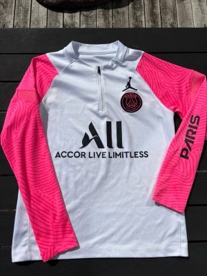 PSG träningströja vit/rosa Jordan - Säljer en långärmad PSG träningströja från Jordan i vitt med rosa mönstrade ärmar. Tröjan har halv dragkedja, svart PSG-logga och Jordan-logga på bröstet samt texten 'PARIS' på ena ärmen. Tillverkad i ett lätt och funktionellt material.