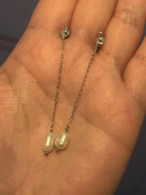 Örhängen med vita sötvattenspärla liknar Edblad  - Säljer ett par stilrena örhängen i stål silverfärgad metall med tunna kedjor och vita söttvattenpärlor längst ner. Örhängena har minimalistisk design och ger en elegant touch till din look.