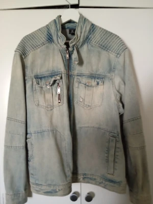 Jeansjacka SMOG - Säljer en ljusblå jeansjacka med coola detaljer som ribbade axlar, bröstfickor med knapp och dragkedja samt ståkrage. Jackan har även sidofickor och en snygg tvättad look. Perfekt för dig som vill ha en unik denimjacka med edge.