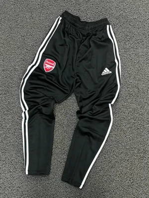 Svarta Arsenal träningsbyxor Adidas - Svarta träningsbyxor från Adidas med Arsenal-logga på ena benet och vita ränder längs sidorna. Byxorna har smal passform och resår i midjan. Perfekta för fotbollsträning eller gymmet.