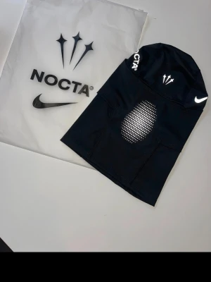 Svart balaclava Nike NOCTA - Svart balaclava från Nike NOCTA med vita loggor och prickdetaljer vid munnen. Tillverkad i stretchigt, tekniskt material som andas. Snygg och sportig design med NOCTA- och Nike-symboler på flera ställen.