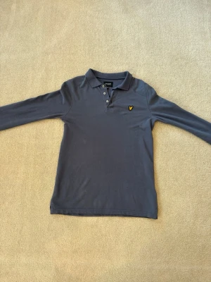 Grå/blå långärmad piké från Lyle & Scott - Snygg grå långärmad pikétröja från Lyle & Scott med klassisk krage och knappar framtill. Tröjan har det ikoniska gula logomärket på bröstet och är tillverkad i mjuk bomull. Perfekt för en stilren och avslappnad look. Köptes för 529kr