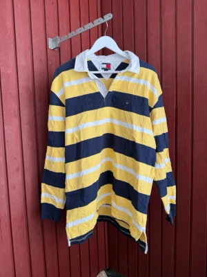 Randig rugbytröja Tommy Hilfiger - Snygg långärmad rugbytröja från Tommy Hilfiger med breda gula, marinblå och vita ränder. Klassisk vit krage och knappslå framtill. Perfekt för en sportig och avslappnad stil. Tröjan har en liten logga på bröstet och är tillverkad i bomull. Lagat hål som syns på bild.