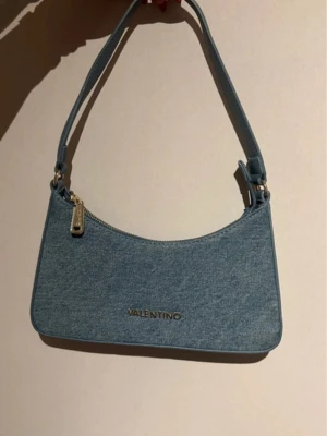 Ljusblå denim handväska Valentino - Trendig ljusblå handväska från Valentino i denimtyg med guldfärgad dragkedja och logga framtill. Väskan har en enkel axelrem. Aldrig använd så helt ny! 