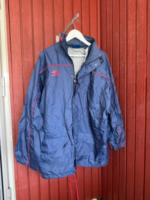 Blå vindjacka från Umbro med röda detaljer - Snygg blå vindjacka från Umbro med röda detaljer och logga på bröstet. Jackan har dragkedja, tryckknappar, två stora fickor framtill och justerbar huva. Perfekt för blåsiga dagar och sportiga outfits.
