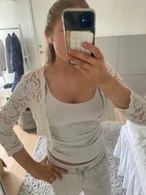 Vit spetsbolero med trekvartsärm - Söt vit bolero i spets med trekvartsärmar och öppen framtill. Perfekt att slänga över ett linne eller klänning för en extra touch. Spetsen har ett blommigt mönster och plagget är kort i modellen.