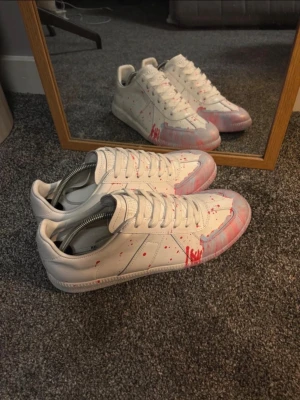 Vita sneakers med röd färgstänk - Unika vita maison margiella skor med coola röda färgstänk och detaljer i rosa på tån. Skorna har klassisk låg profil, vita snören och är tillverkade i läder. Perfekta för dig som vill sticka ut med en kreativ och modern stil.