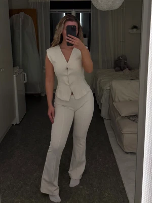 Krävit set med väst och byxor - Säljer ett stilrent set i krämvit färg bestående av en figursydd väst med knappar och matchande höga, utsvängda byxor. Materialet ser ut att vara stretchigt och bekvämt, perfekt för dig som gillar en clean och modern look.
