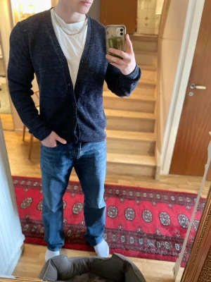 Mörkblå cardigan - Mörkblå cardigan med två knappar framtill och diskret mönster i tyget. Tröjan har v-ringad halsringning och ribbade muddar vid ärmslut och nederkant. Perfekt att slänga över en t-shirt för en avslappnad look.